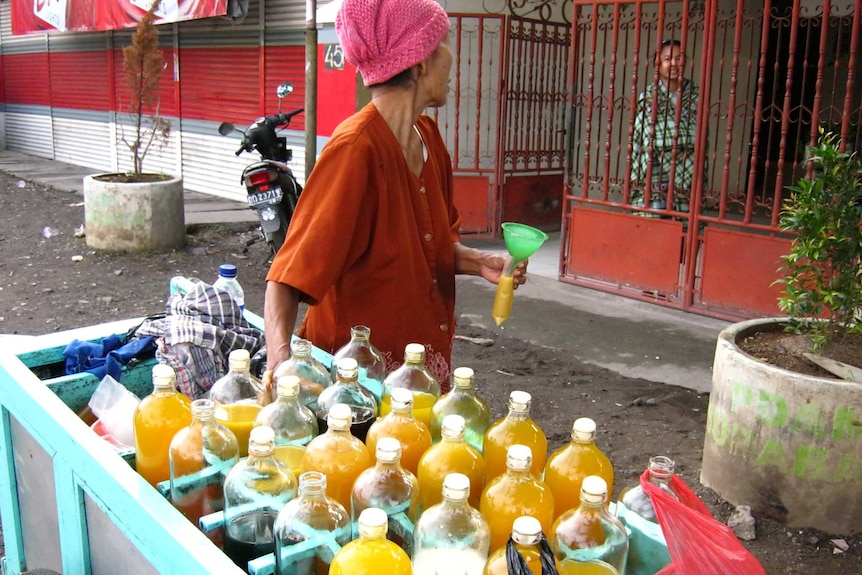 An Indonesian woman looks over her shoulder as she holds a bag of jamu and rests her hand on a jamu cart.