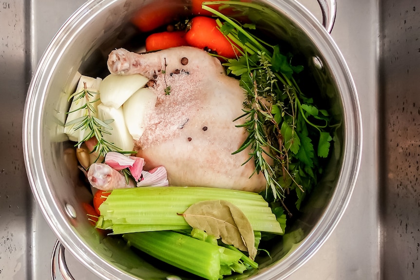 A whole chicken in a stock pot with celery, carrots, garlic and herbs, ready for poaching.
