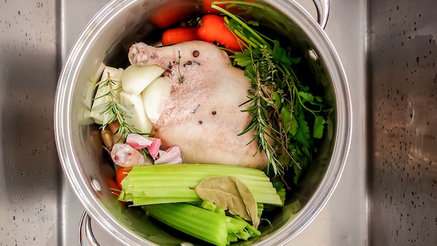 A whole chicken in a stock pot with celery, carrots, garlic and herbs, ready for poaching.