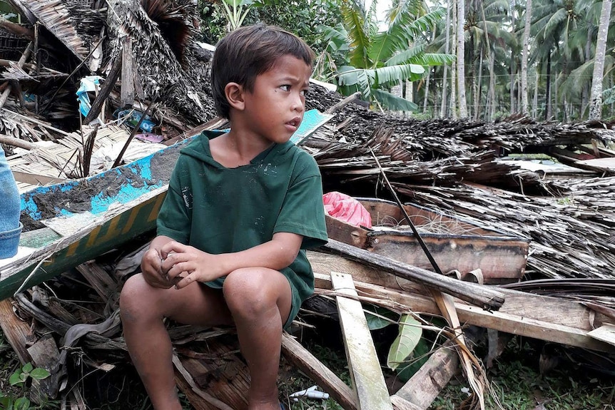 A young Filipino boy sits on planks of wood of a destroyed hut with palm trees in the background.