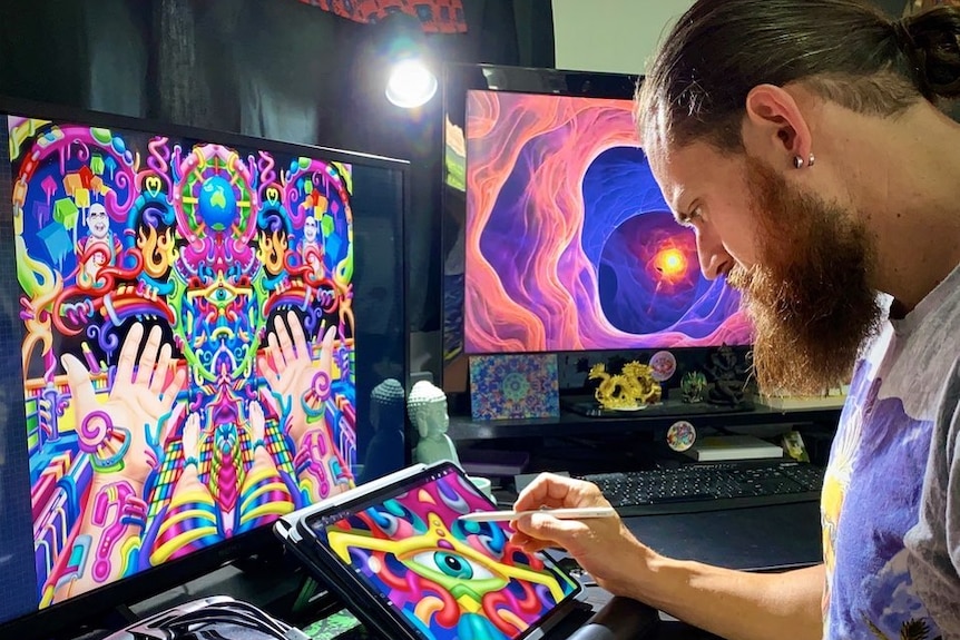 Ayjay uses a stylus on a small screen showing a colourful digital artwork, near other artworks on bigger screens. 