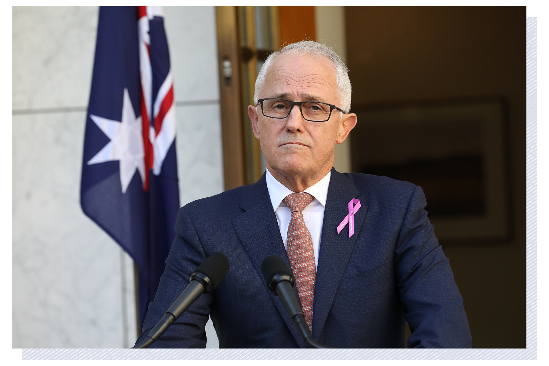 Dressed in a suit with a purple ribbon on his lapel, Malcolm Turnbull stands at a lectern, looking serious.