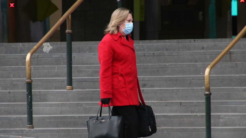 A woman wearing a red coat and surgical mask, who is carrying a computer bag, walks past Flinders Street Station.