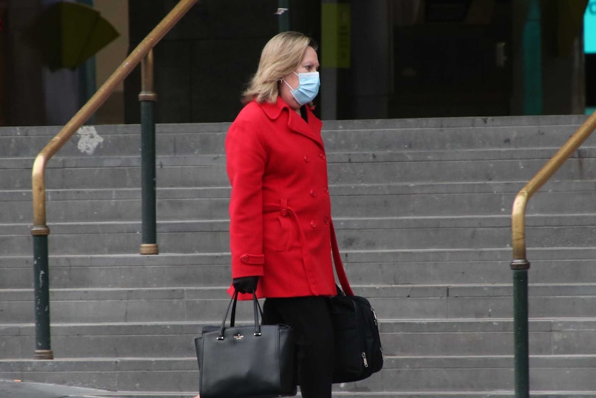 A woman wearing a red coat and surgical mask, who is carrying a computer bag, walks past Flinders Street Station.