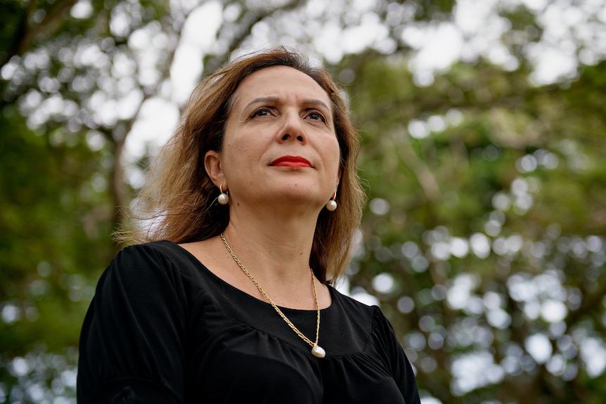 A woman wearing a black top stands in a park and looks to the distance. She has a peaceful expression.