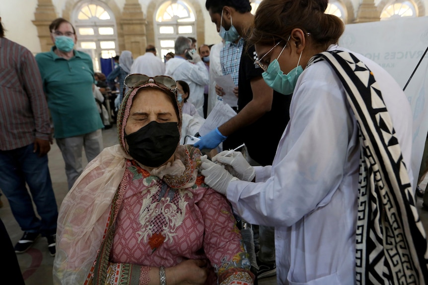 A woman wearing a pink headscarf and mask reacts while receiving a Sinopharm coronavirus vaccine from a health worker.