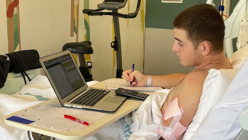 A boy with his injured left arm bandaged looking at a laptop while doing homework in a hospital bed