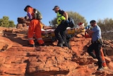 Six people in police and SES uniforms carry a stretcher up the side of a red rockface