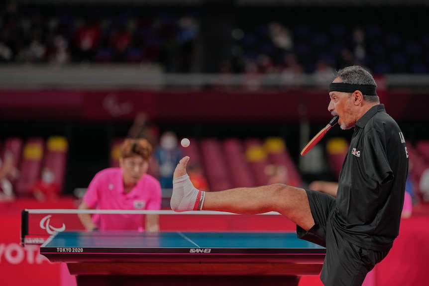A double amputee Paralympic table tennis player flicks ball up with his foot as he holds paddle in his mouth ready to serve..