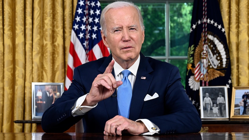 Joe Biden sits at a desk it he Oval Office of the White House and addresses the camera