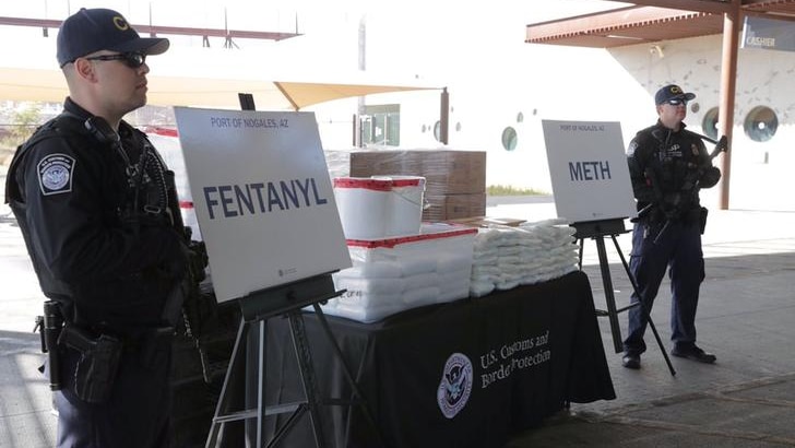 Two officers in black shirts, trousers and caps hold guns and stare ahead. A table with fentanyl and meth packets between them.