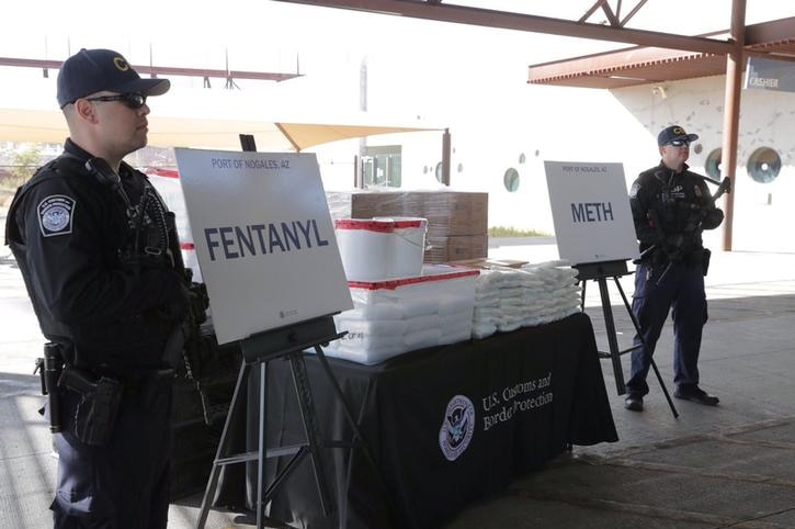 Two officers in black shirts, trousers and caps hold guns and stare ahead. A table with fentanyl and meth packets between them.