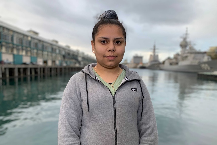 A young woman stands in front of a body of water and Navy ships.