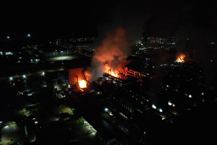 An aerial view of a factory complex at night, with fires glowing from within buildings.