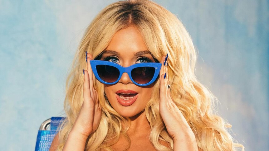A blonde Megan Moroney wears a blue polka dot dress and blue sunglasses. Her face has a surprised expression.
