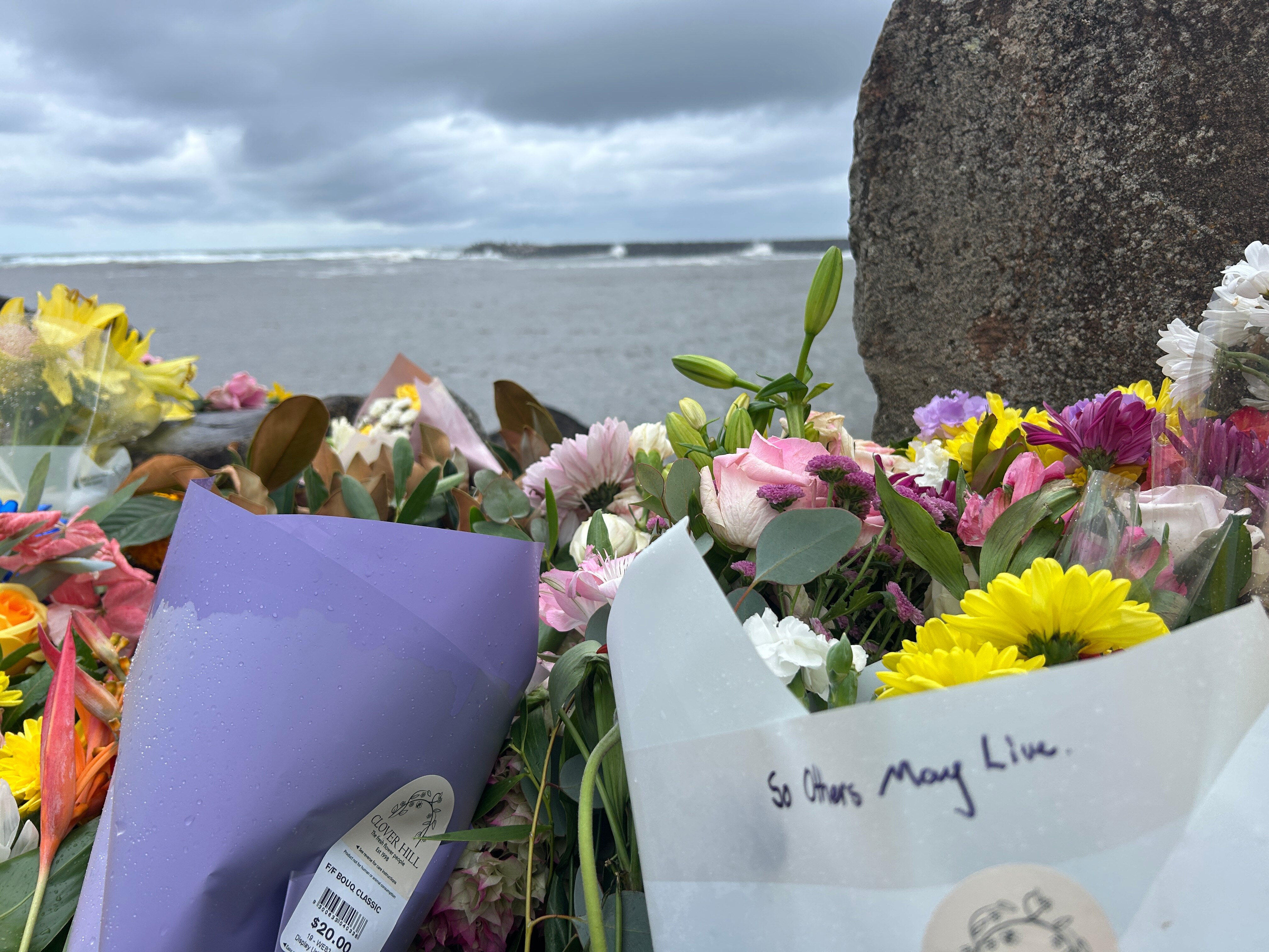 Floral tribute with message 'So others may live' and river and breakwall in background.