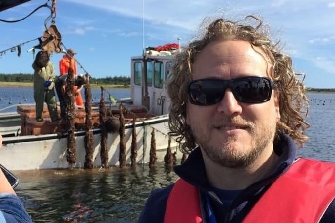 Shane Roberts wears sunglasses and a lifejacket on a boat, in the background two men farm pull mussels out of water