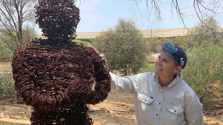 A woman is smiling, looking at a sculpture of a person made out of rusty barbed wire. She has her hand on its shoulder.