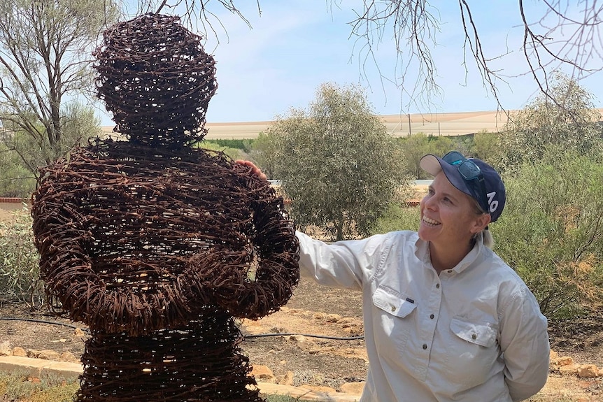 A woman is smiling, looking at a sculpture of a person made out of rusty barbed wire. She has her hand on its shoulder.