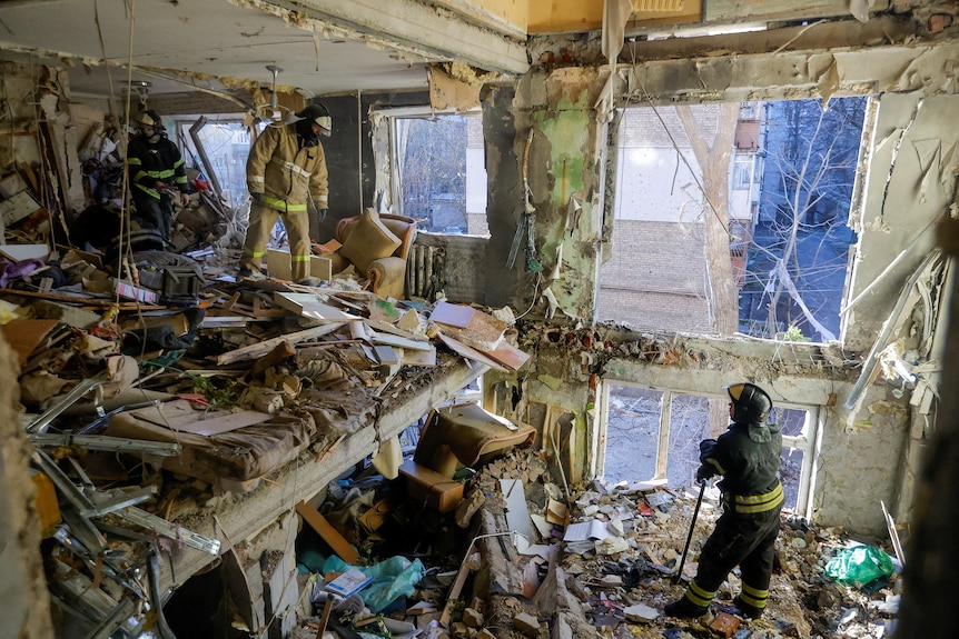 Two men stand inside an apartment that has been destroyed by shelling with debris and destruction everywhere. 