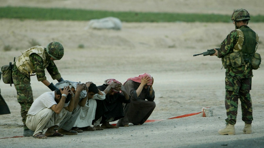 Five Iraqi men squat on the ground with their hands on their heads. Soldiers with guns stand over them