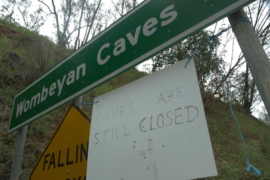 A hand written sign attached to a road sign that says the Wombeyan Caves are still closed.