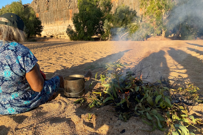 An older woman sits by a small fire on the sand in Danggu Gorge.