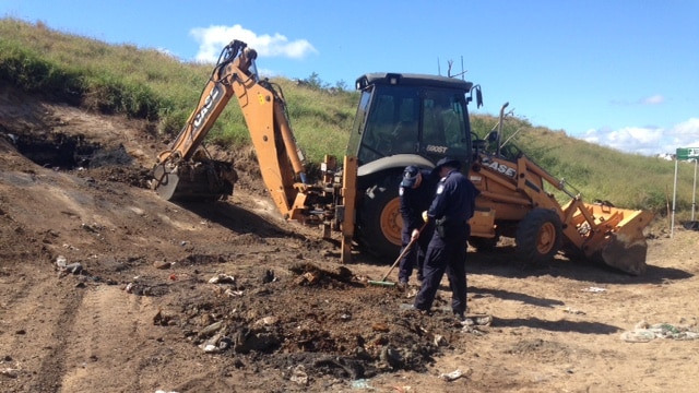A front-end loader is digging up dirt at a dump site while a police officer rakes through debris.