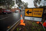 A sign saying COVID-19 testing on a road with cars parked next to it