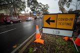 A sign saying COVID-19 testing on a road with cars parked next to it