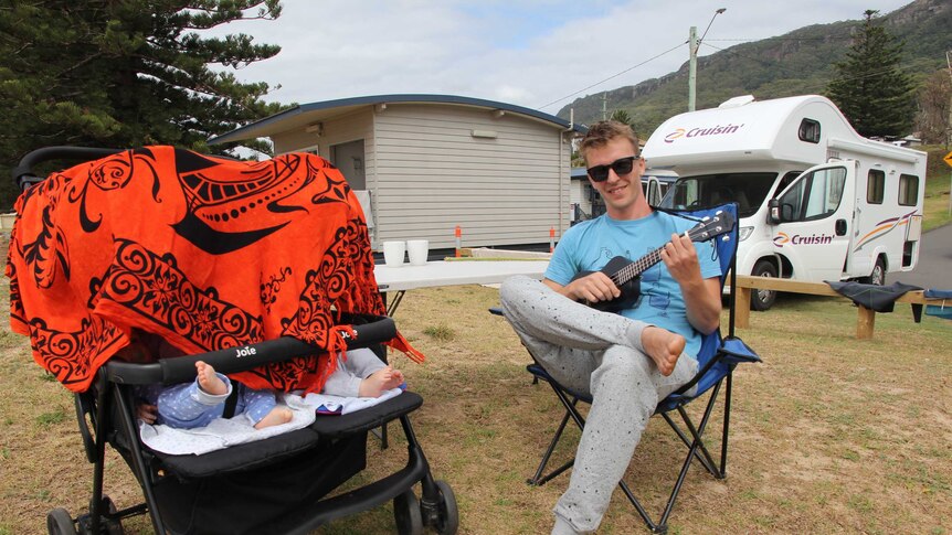 Camper traveller Bernt Haupt relaxes in a chair with his ukelele in front of his campervan, next to a pram with his twin boys
