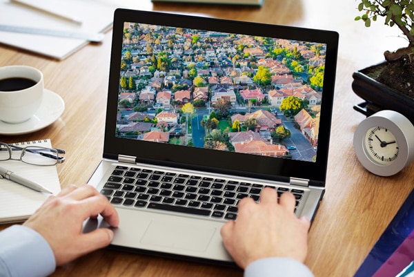 A laptop computer sits on a table showing houses on its screen with a man's hands resting on the keyboard.