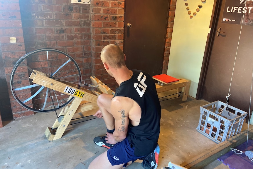 Matthew Nunan kneeling in front of a homemade rowing machine, made of timber and a bike wheel.