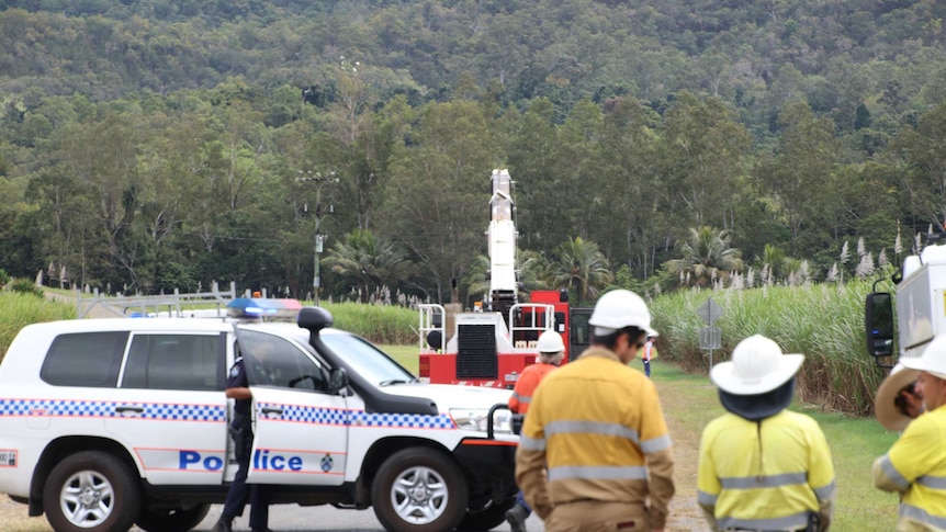 Work crews stand behind a police car to look at a crane next to a sugar field and powerlines