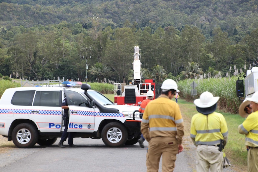 Work crews stand behind a police car to look at a crane next to a sugar field and powerlines