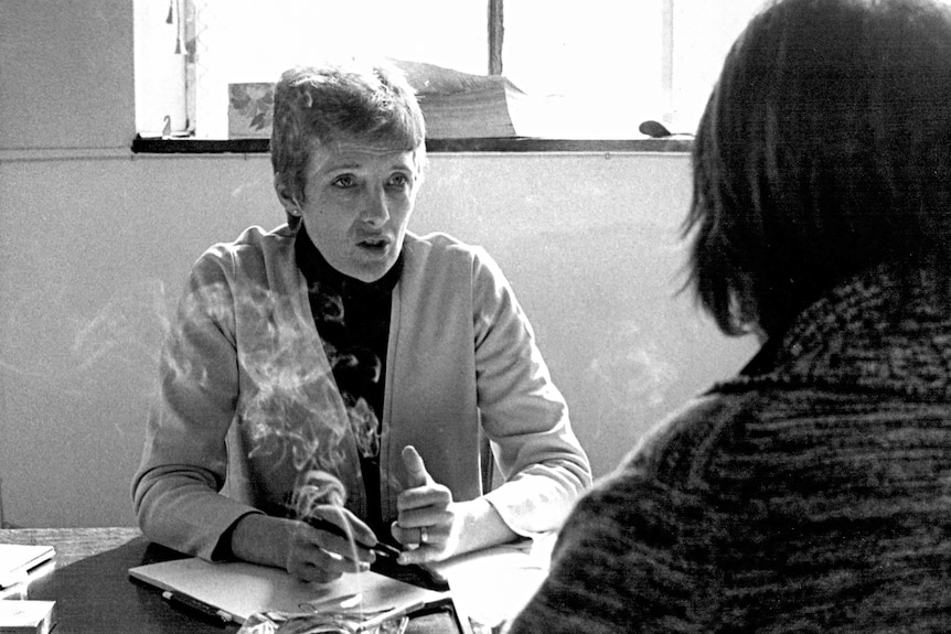 A woman with short hair sitting at a desk and smoking a cigarette while talking to someone sitting opposite.