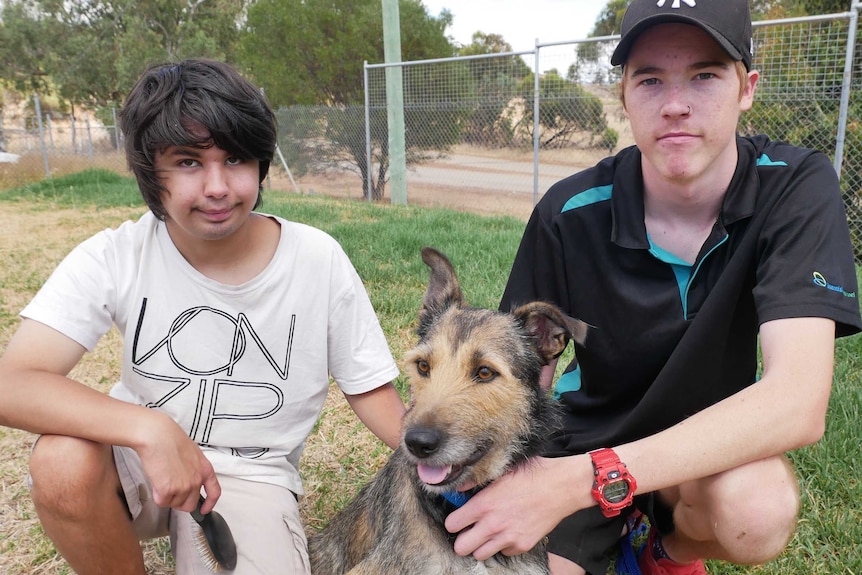 A boy in a white shirt crouches next to a dog with a young man in a black shirt and hat alongside them.