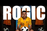 Tom Rogic holds a ball and stares at the camera in his Socceroo uniform, with his name in large letters behind him.