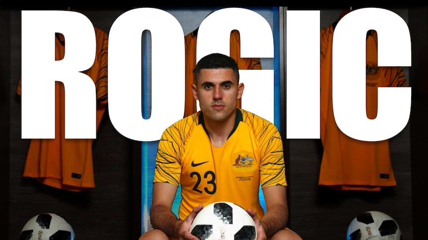 Tom Rogic holds a ball and stares at the camera in his Socceroo uniform, with his name in large letters behind him.