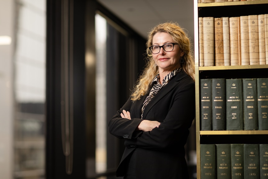 A woman with long blonde hair and a black jacket stands with her back leant on a bookshelf. 