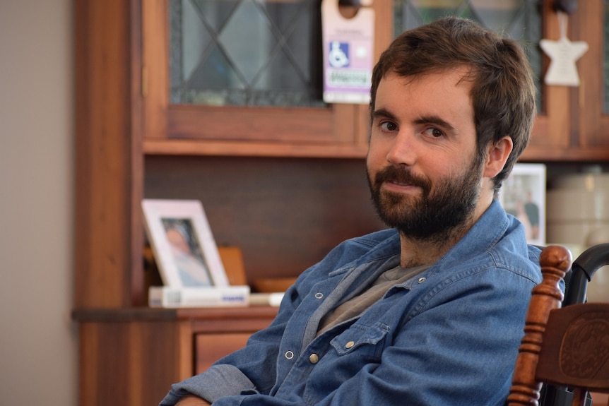 A man sits in a wheelchair smiling at the camera, he has short brown hair and a beard.