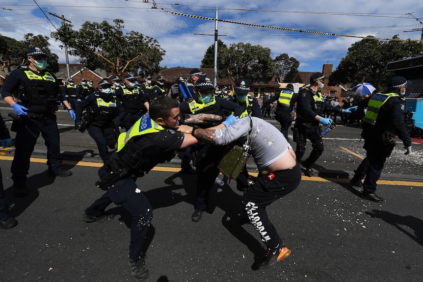 A police officer and protester wrestle, standing, on a suburban road surrounded by other officers.
