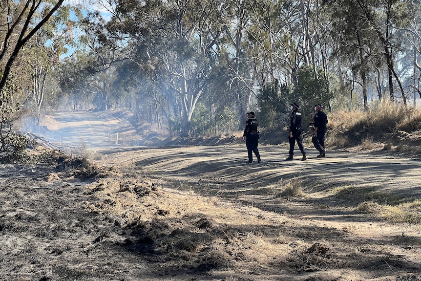 Three police officers walk along a dirt road that has blackened scrub along it.