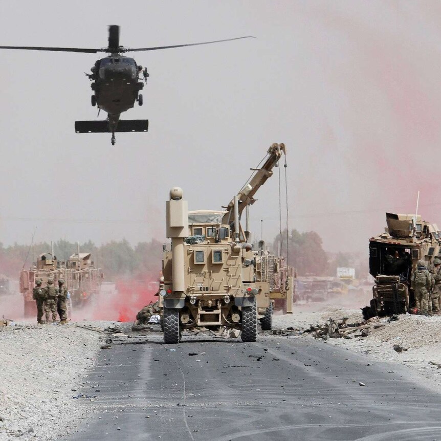 A military helicopter hovers over a road above other heavy machinery next to a damaged military vehicle.