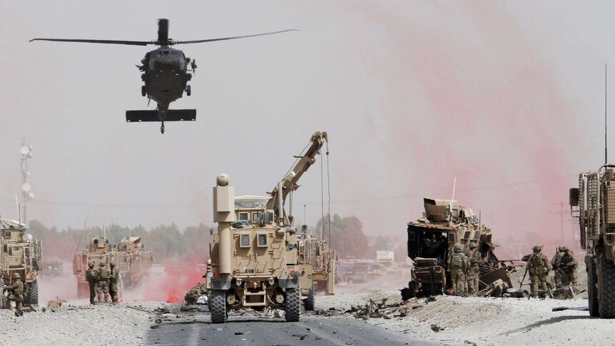 A military helicopter hovers over a road above other heavy machinery next to a damaged military vehicle.