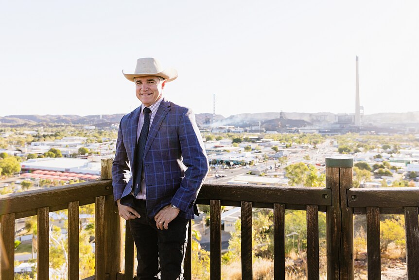 A man wearing a suit jacket and a cowboy hat stands at a lookout with mining stacks in the background