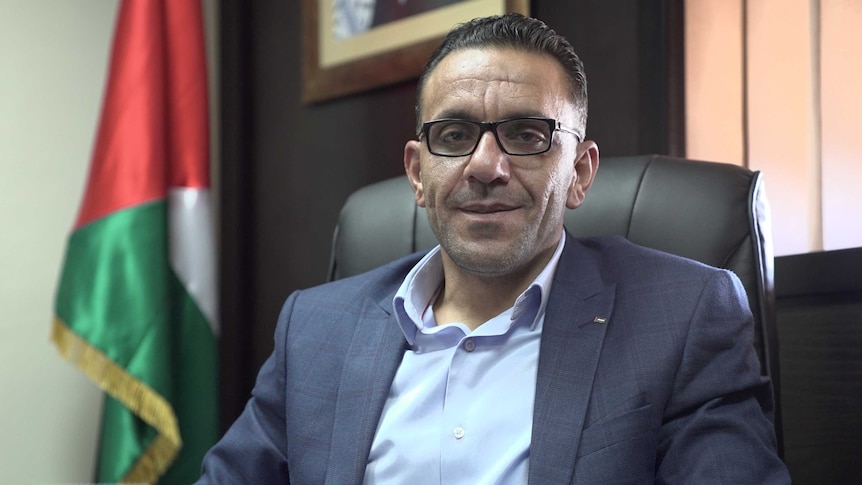 Adnan Gaith sits at a desk, wearing a suit, with a neutral facial expression.