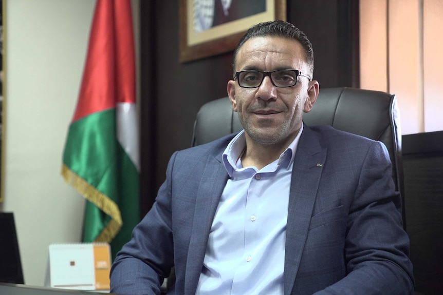 Adnan Gaith sits at a desk, wearing a suit, with a neutral facial expression.