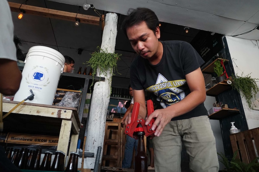 A man leans over a beer bottle with a red device as he and another man out of shot bottle beer