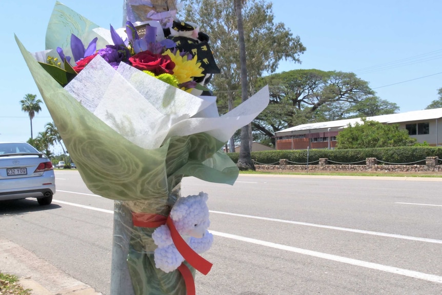 Colourful flowers and a small white teddy are taped to a light pole, next to a road